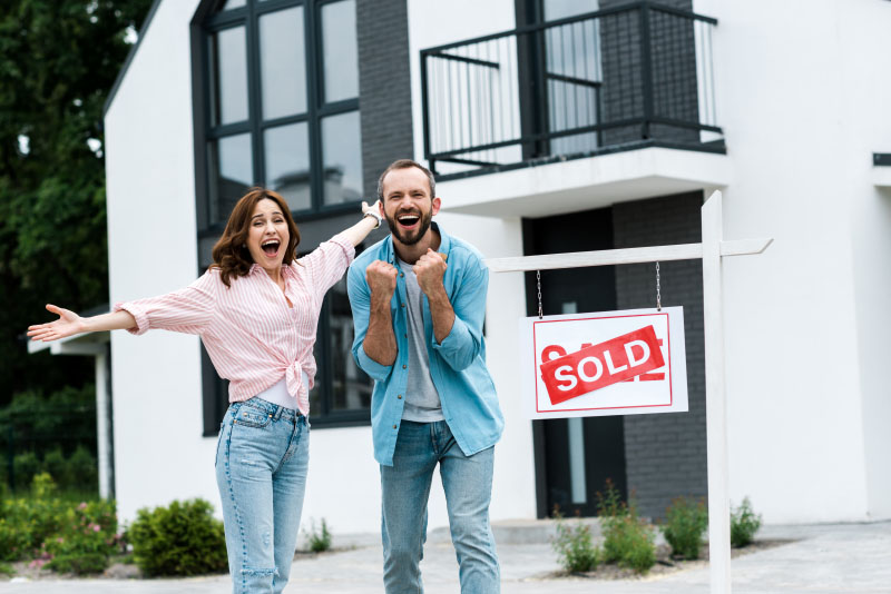 an excited man and woman standing next to a sold sign in front of their house
